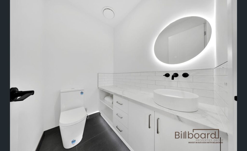 Contemporary bathroom featuring a white toilet, sink, and illuminated round mirror on a tiled wall.