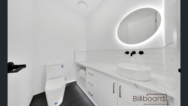 Contemporary bathroom featuring a white toilet, sink, and illuminated round mirror on a tiled wall.