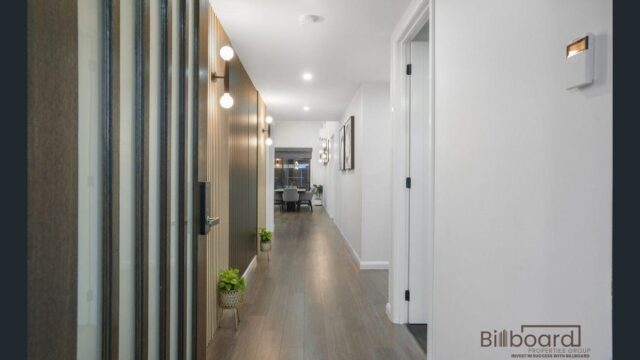 Long modern hallway with timber flooring, vertical wood feature wall panels, decorative wall lights, white side walls, and a view leading toward the dining area at the end of the corridor.