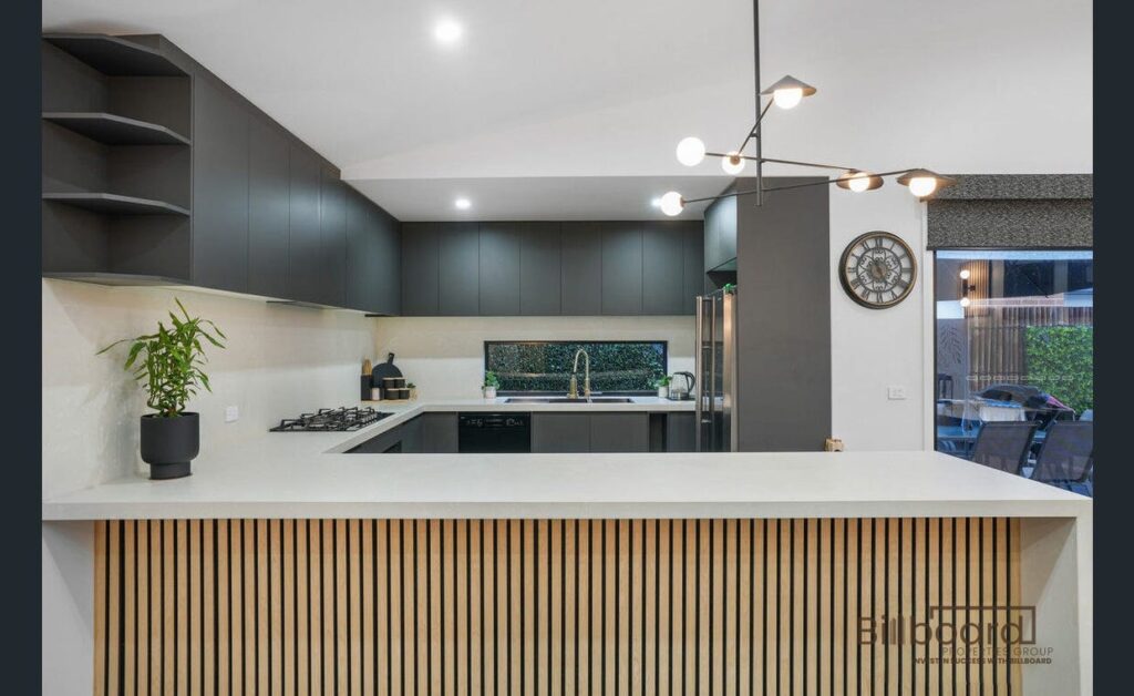 Contemporary kitchen featuring a sleek island bench with vertical timber slat detailing, matte dark cabinetry, built-in gas cooktop, modern pendant lighting, stainless steel appliances, and a large window splashback overlooking greenery outside.