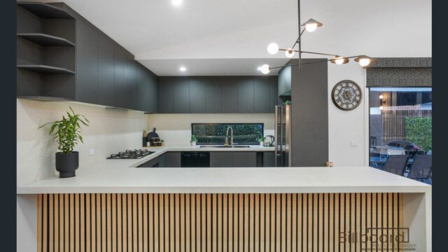 Contemporary kitchen featuring a sleek island bench with vertical timber slat detailing, matte dark cabinetry, built-in gas cooktop, modern pendant lighting, stainless steel appliances, and a large window splashback overlooking greenery outside.