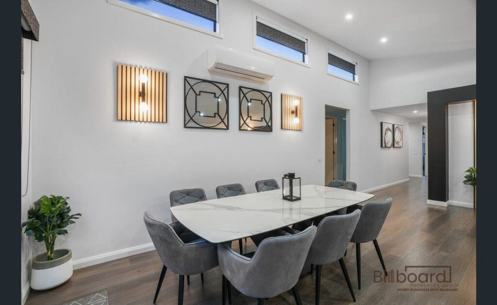 Modern dining room with a large marble-style dining table surrounded by grey upholstered chairs, wall-mounted decorative lighting, artwork pieces, high horizontal windows providing natural light, and wooden flooring for a contemporary interior look.