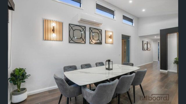 Modern dining room with a large marble-style dining table surrounded by grey upholstered chairs, wall-mounted decorative lighting, artwork pieces, high horizontal windows providing natural light, and wooden flooring for a contemporary interior look.