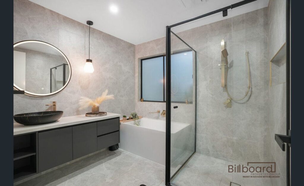 Luxurious modern bathroom with grey stone textured walls, a matte black vanity and round vessel sink, hanging pendant light, built-in bathtub under a frosted window, and a glass-framed walk-in shower featuring brushed gold fixtures and rain shower head.