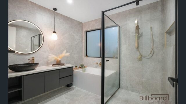 Luxurious modern bathroom with grey stone textured walls, a matte black vanity and round vessel sink, hanging pendant light, built-in bathtub under a frosted window, and a glass-framed walk-in shower featuring brushed gold fixtures and rain shower head.