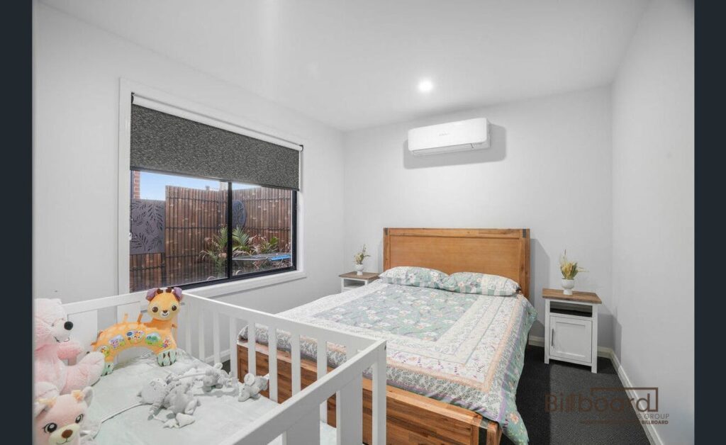 Bright bedroom with a wooden double bed, a matching bedside table, wall-mounted air conditioner above the bed, and a white baby cot placed beside the bed filled with plush toys. The room has a large window with a roller blind and simple modern decor.