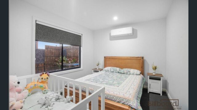 Bright bedroom with a wooden double bed, a matching bedside table, wall-mounted air conditioner above the bed, and a white baby cot placed beside the bed filled with plush toys. The room has a large window with a roller blind and simple modern decor.