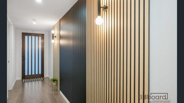 Stylish hallway with modern vertical timber feature wall panels in contrasting light and dark tones, warm wall lights, timber flooring, and a glass-panel front door at the end of the corridor.