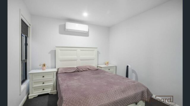 Cozy bedroom with a white bed frame, matching white side tables, neatly made bed with a mauve quilt, small decorative items on the bedside tables, and a wall-mounted air conditioner above the headboard. The room has simple modern decor with minimal furnishings.