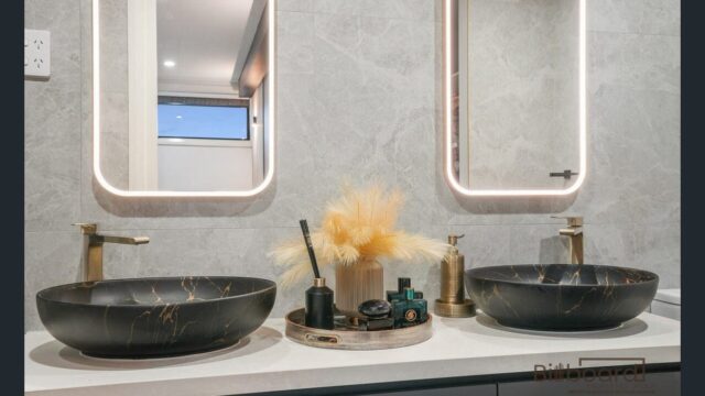 Modern bathroom vanity with two matte black marble-style vessel sinks, gold tapware, decorative tray with accessories placed between the sinks, and two illuminated rectangular mirrors mounted on a grey stone-textured wall.