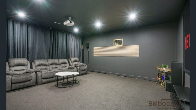 Modern home theatre room featuring a row of reclining cinema-style seats, dark carpet flooring, black walls with acoustic paneling, ceiling-mounted projector, round coffee table, and a small side bar setup in the corner, creating a cozy cinema-like environment.