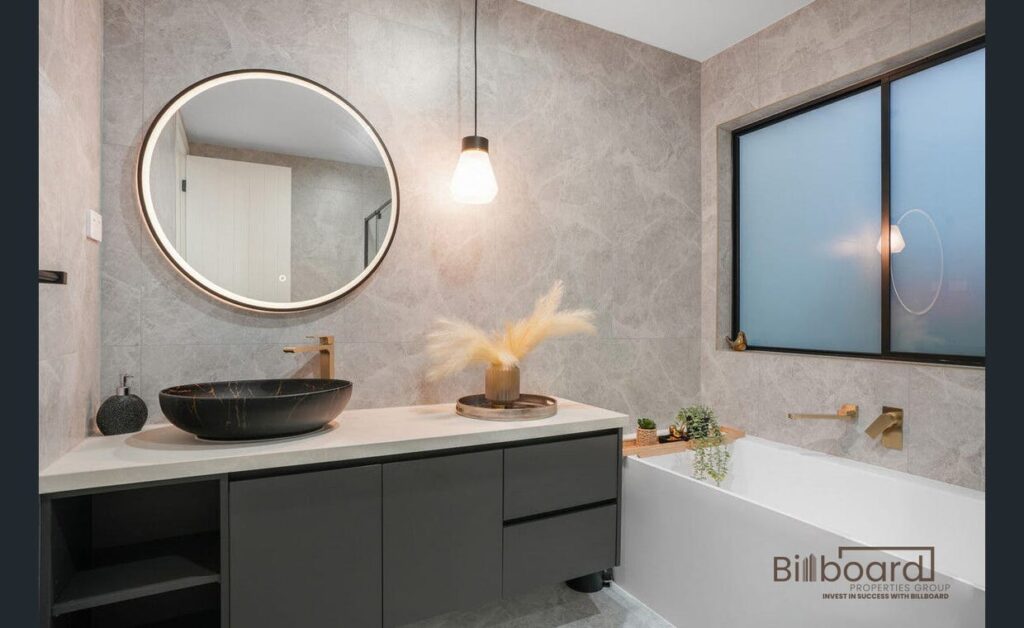 Stylish modern bathroom featuring a matte black vanity with a round vessel sink, large round mirror, hanging pendant light, grey stone-textured walls, built-in white bathtub with brushed gold tapware, and a frosted black-framed window adding privacy and contemporary elegance.