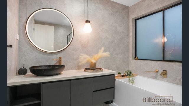 Stylish modern bathroom featuring a matte black vanity with a round vessel sink, large round mirror, hanging pendant light, grey stone-textured walls, built-in white bathtub with brushed gold tapware, and a frosted black-framed window adding privacy and contemporary elegance.