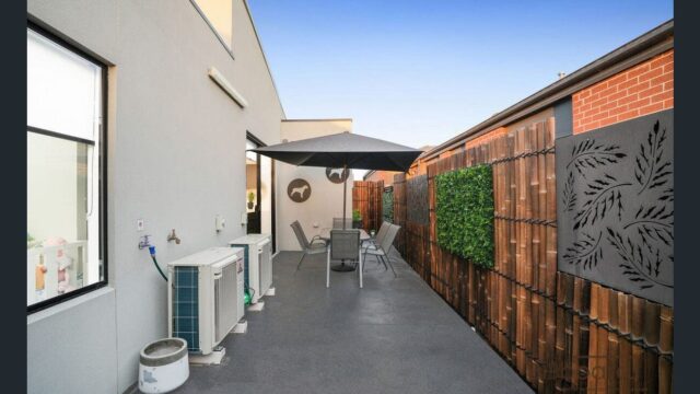 Outdoor patio area with paved flooring, outdoor seating under a large umbrella, air conditioning unit along the wall, and a decorative wooden fence with greenery and metal art panels on one side. The space is narrow and neatly maintained with a modern backyard setup.