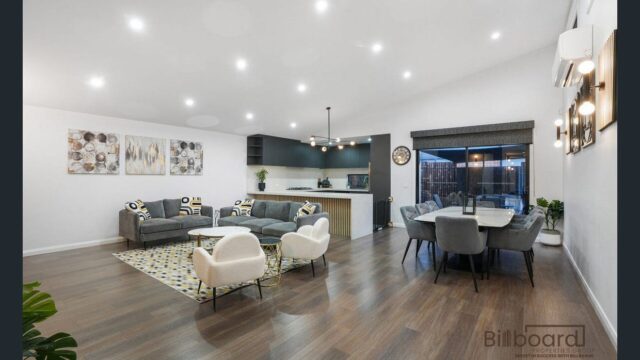 Spacious open-plan living and dining area with modern grey sofas, white accent chairs, round coffee tables, patterned rug, large dining table with grey chairs, timber flooring and a contemporary kitchen with dark cabinetry and timber feature island bench in the background.
