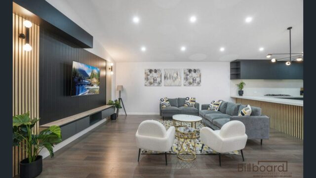 Modern living room with timber flooring, feature wall with mounted TV and vertical timber paneling, stylish round coffee tables, grey sectional sofa, two white accent armchairs, and open view to the kitchen area in the background with contemporary lighting and decor.