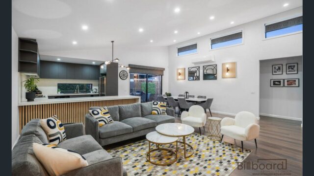 Open plan living and dining area featuring modern grey sofas, accent chairs, patterned rug, round coffee tables, stylish feature lighting, and a contemporary kitchen in the background with timber slat island bench and dark cabinetry. The space is bright with multiple high windows and wooden flooring throughout.