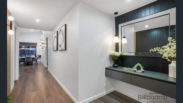Contemporary hallway nook with a modern floating console table, decorative flowers, pendant wall lights, and a large square mirror framed by vertical timber paneling, with timber flooring extending through the corridor leading to the dining area.