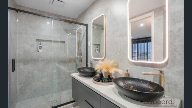 Modern bathroom with dual matte black vessel sinks on a sleek vanity, illuminated rectangular mirrors, brushed gold tapware, decorative tray with styling items, and a glass enclosed walk-in shower with grey stone textured walls.